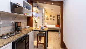 A compact kitchen with a stove, microwave, and coffee maker. A small dining area with a table and chairs is visible. The room has a neutral color scheme and a view into the living area with a bed and a wall decorated with framed pictures.