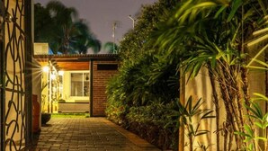 A nighttime view of a house with a paved driveway leading to a glass door entrance, surrounded by lush greenery and a brick wall.