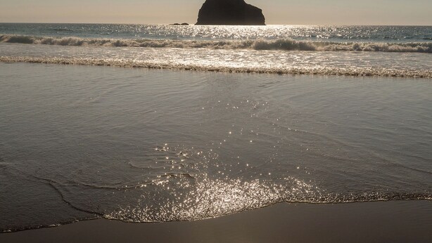 Une plage où les vagues se brisent près du rivage, avec une formation rocheuse visible au loin.