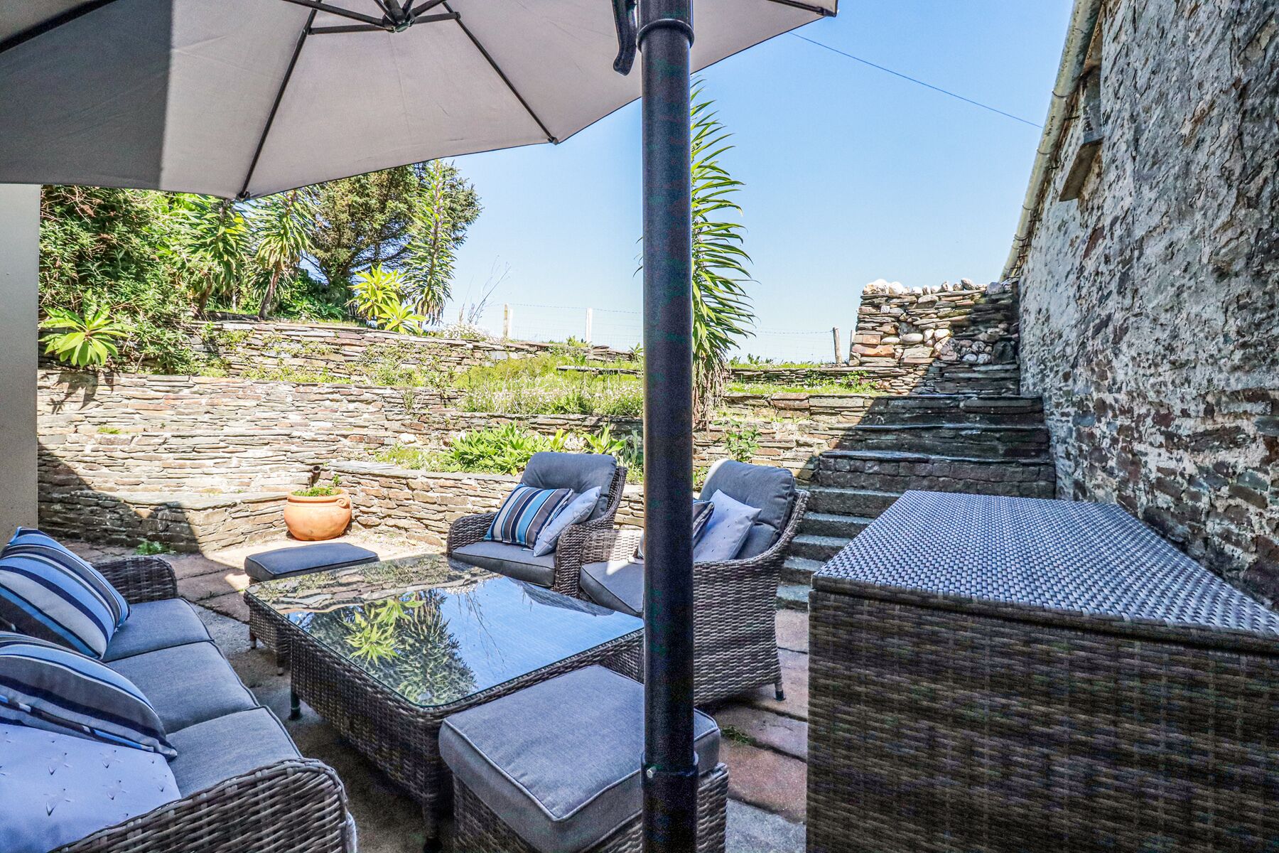 An outdoor seating area with wicker furniture and a glass-top table, a stone wall, and a view of a natural landscape.