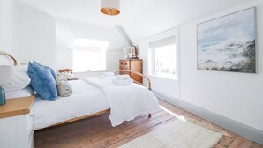 Une chambre à coucher avec un lit en bois, du linge de lit blanc, une commode en bois, une table de chevet, une lampe et un tableau représentant une scène de plage.