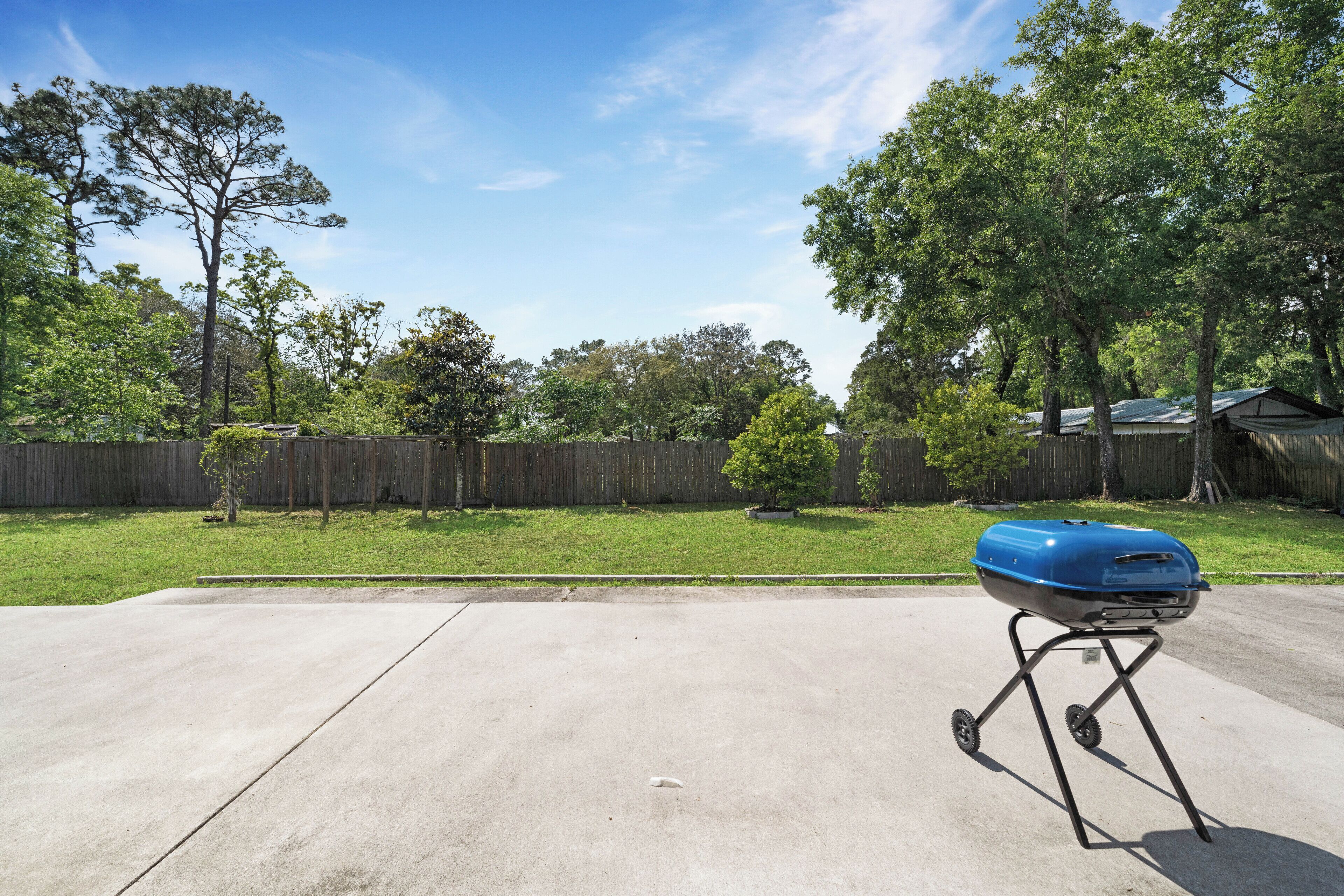 A backyard with a concrete patio, a blue grill on a portable stand, a wooden fence, and trees in the background.