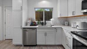 A modern kitchen with white cabinetry, stainless steel appliances, and a window with a view of greenery outside. - Multi Home - Up to 16 people - Balcony Access (Austin)