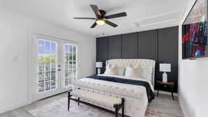 A bedroom with a tufted headboard, a bed with white and dark blue bedding, a bench at the foot of the bed, a ceiling fan, and a TV mounted on the wall. - Multi Home - Up to 16 people - Balcony Access (Austin)
