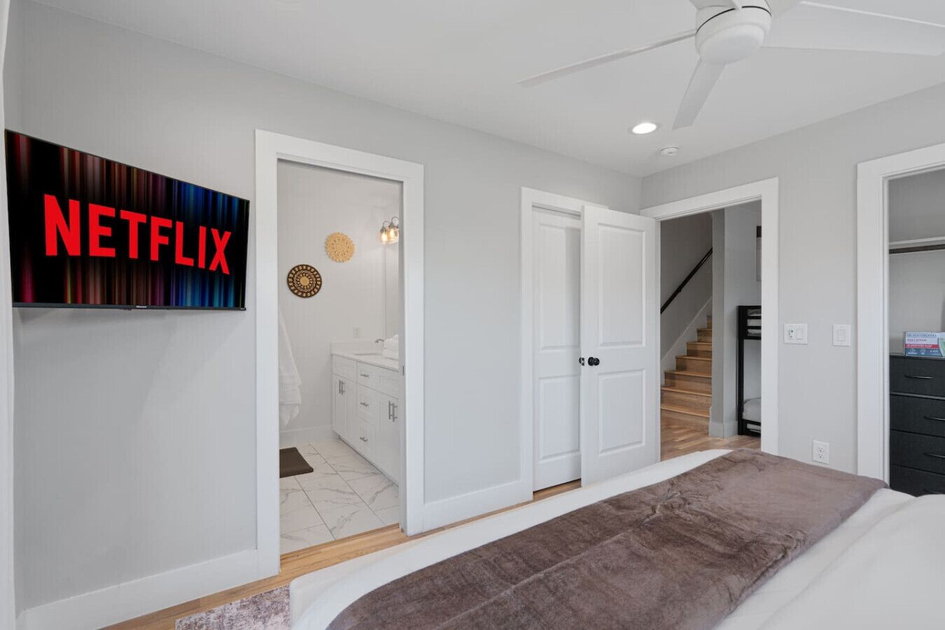 A modern bedroom with a large bed, a ceiling fan, and a wall-mounted TV displaying the Netflix logo.