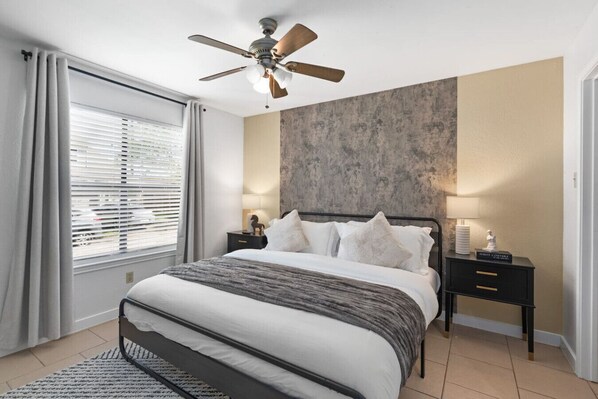 A modern bedroom with a bed, a ceiling fan, a nightstand with a lamp, and a window with blinds.