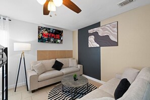 A modern living room with a sofa, a coffee table, and a floor lamp. There is a wall with a Netflix poster and a painting. - Up to 32 People - Luxurious Home (Austin)