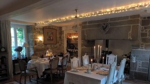 A dining area with a stone fireplace, string lights, and a chandelier. - Le Presbytere (Beauficel)