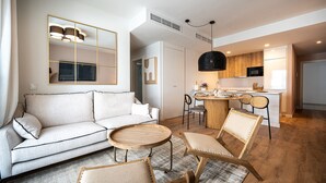 Ein modernes Wohnzimmer mit einer Couch, einem runden Couchtisch und zwei Sesseln. Im Hintergrund ist eine Küche mit Essbereich zu sehen.