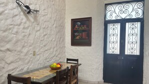 A dining area with a wooden table and chairs, a bowl of fruit, a wall-mounted light fixture, a framed painting, and a black door with decorative glass panels.