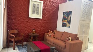 A living room with a red textured wall, a framed artwork, a sofa with patterned cushions, a wooden chair, a small table with a vase, and a trunk with a red cloth.
