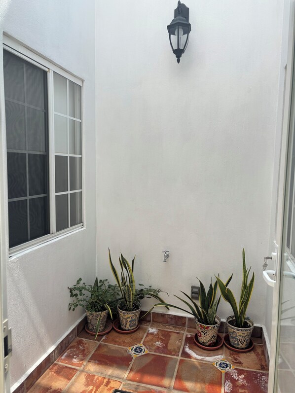Un espacio exterior pequeño y cerrado con una ventana, una lámpara fijada a la pared y varias plantas en macetas.