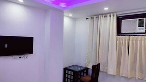 Um quarto pequeno com televisão de tela plana na parede, uma mesinha com uma cadeira e uma janela com cortinas.