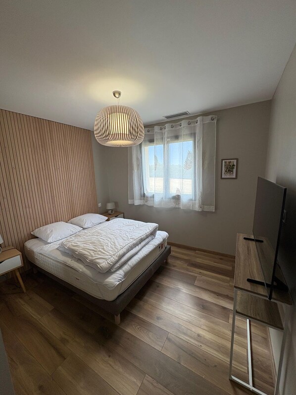 A bedroom with a bed, wooden floor, a TV stand, a window with curtains, and a hanging light fixture.