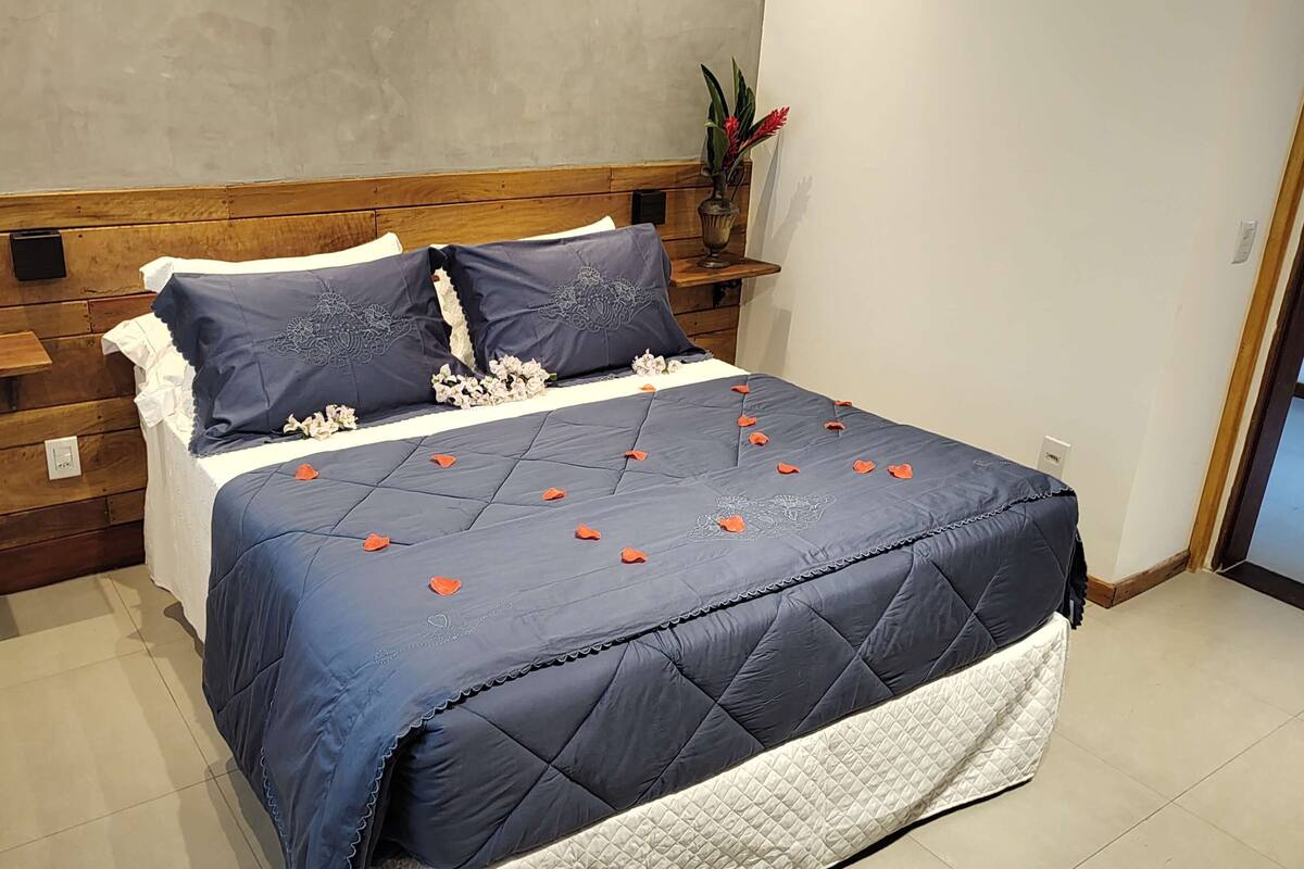 A neatly made bed with a dark blue quilt and decorative pillows, a wooden headboard, and a small potted plant on a nightstand.