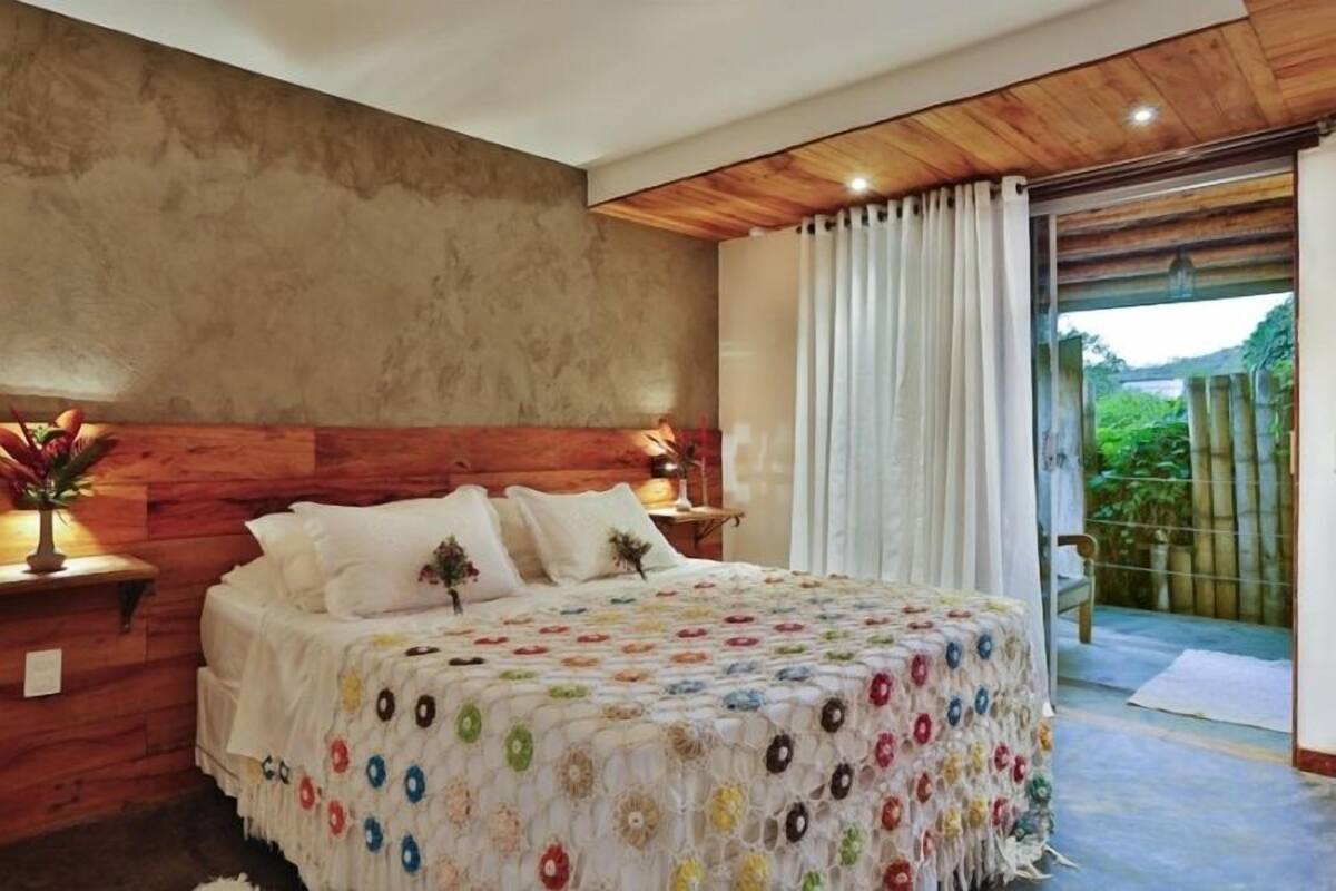 A bedroom with a wooden headboard, a large bed with a colorful floral bedspread, a nightstand with a lamp, and a view of greenery through a sliding glass door.