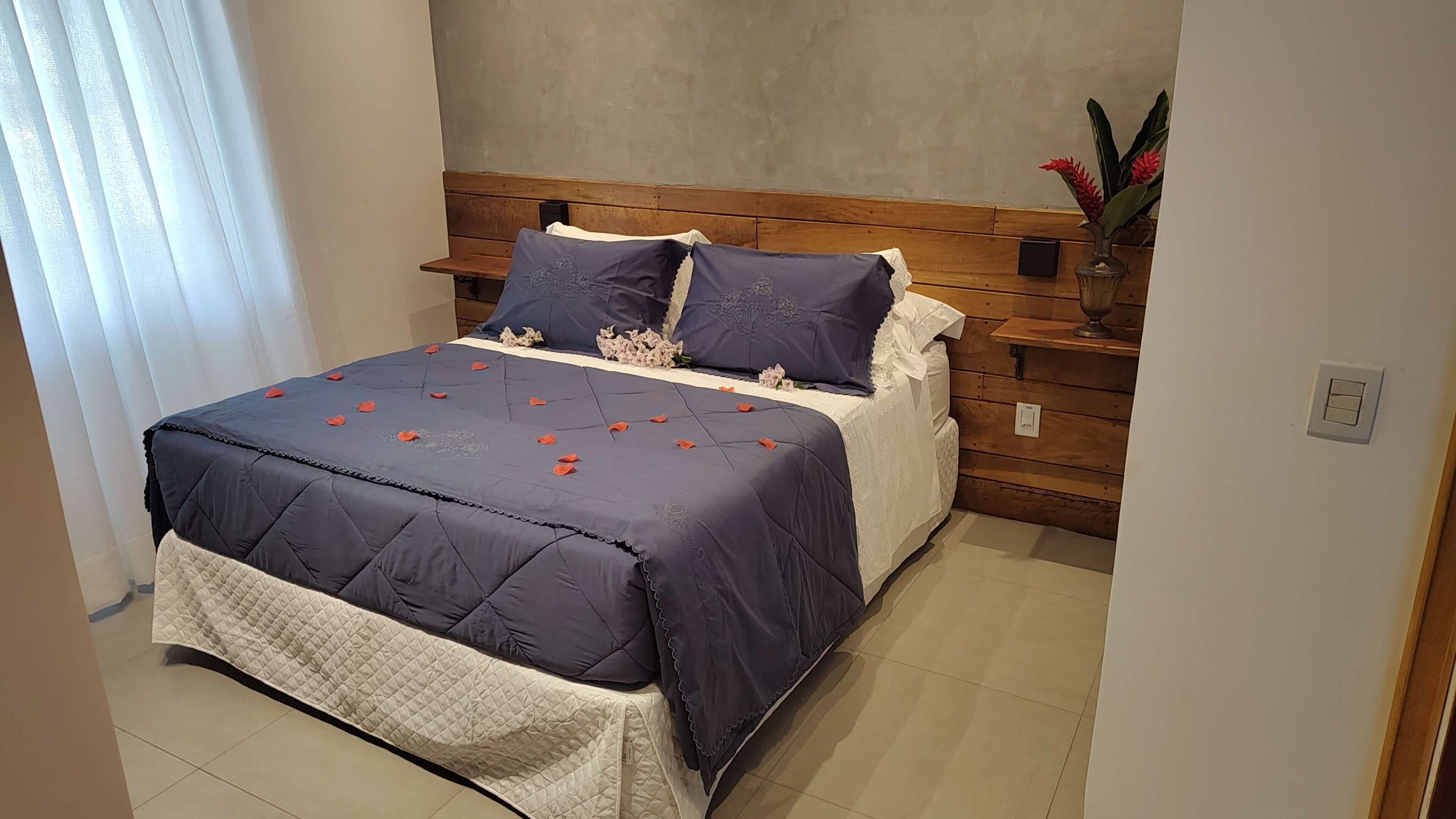 Ein ordentlich bezogenes Bett mit einem dunkelblauen Überwurf und weißer Bettwäsche, ein Holz-Kopfteil und ein kleiner Nachttisch mit einem Blumenstrauß.