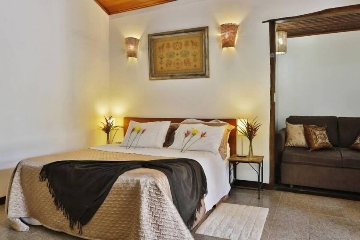 A bedroom with a bed, a nightstand, a sofa, and wall-mounted lighting.