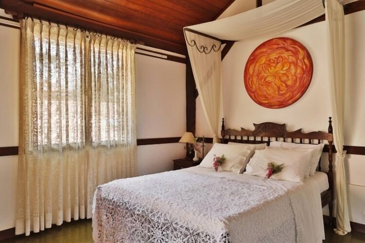A bedroom with a four-poster bed, a large floral artwork on the wall, and a window with sheer curtains.