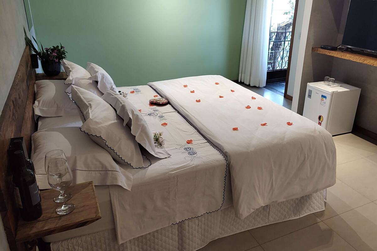 A hotel room with a bed, a bedside table with a wine bottle and glass, a small refrigerator, and a TV on a stand.