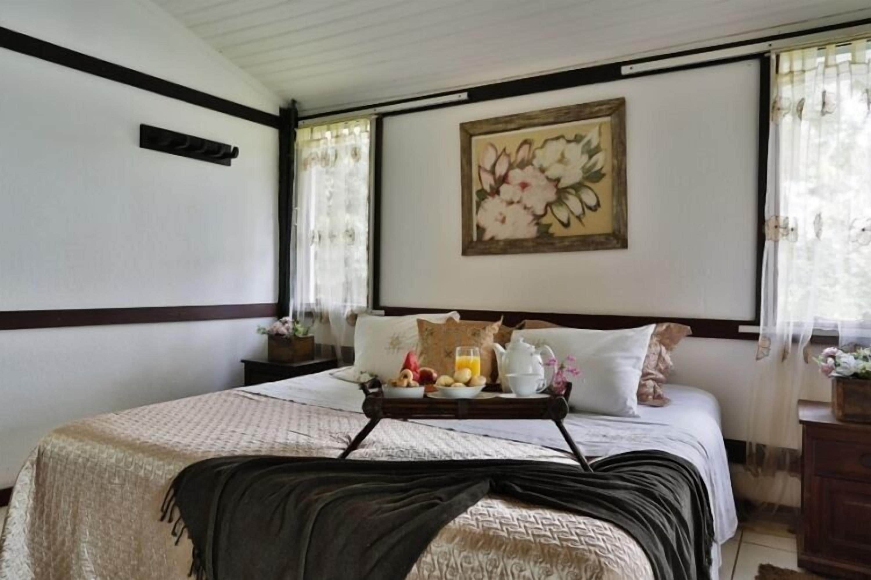 Un lit bien fait, avec un plateau contenant des éléments de petit-déjeuner, un tableau floral accroché au mur et une fenêtre ornée de rideaux légers.