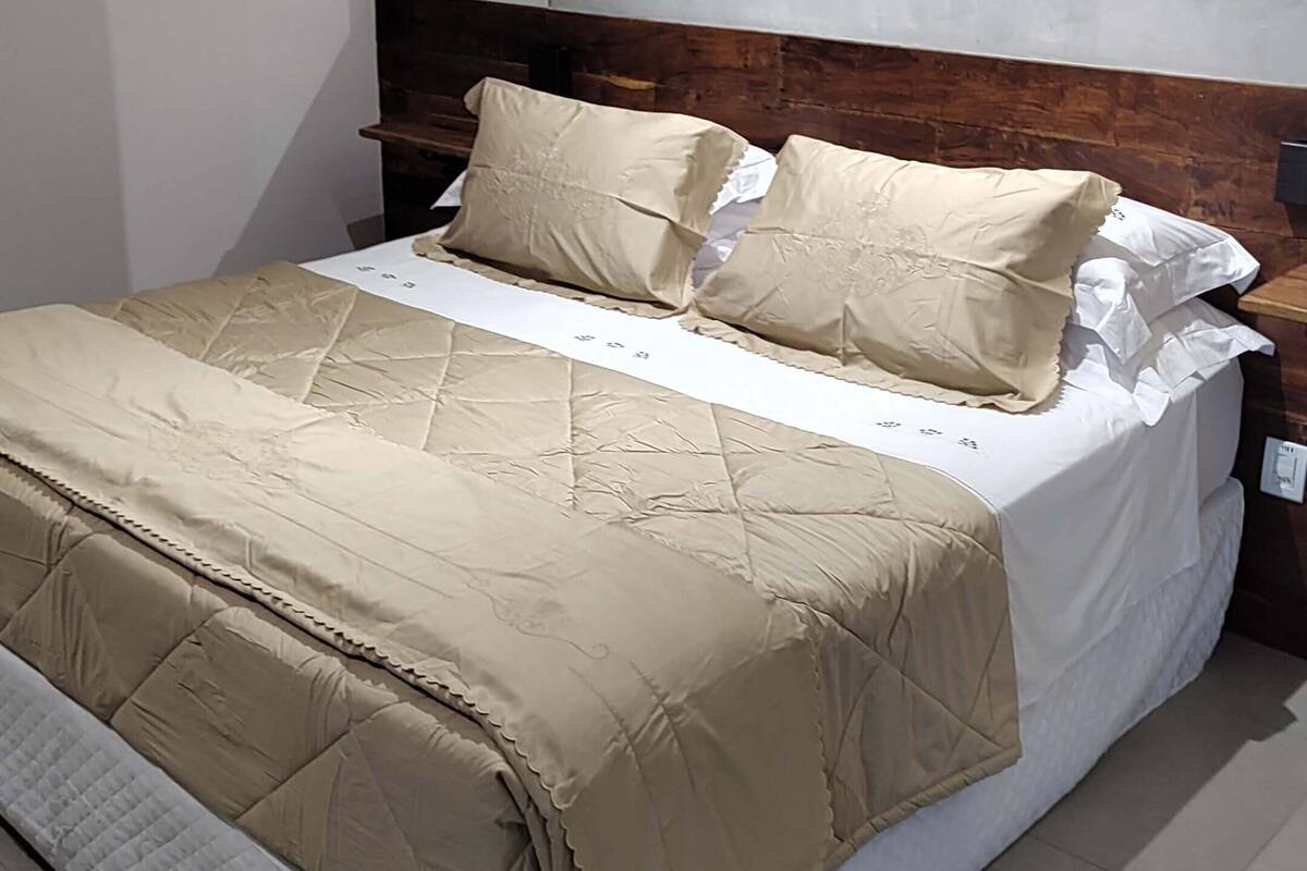 A neatly made bed with a beige quilt and pillows, a wooden headboard, and a wall-mounted air conditioner.