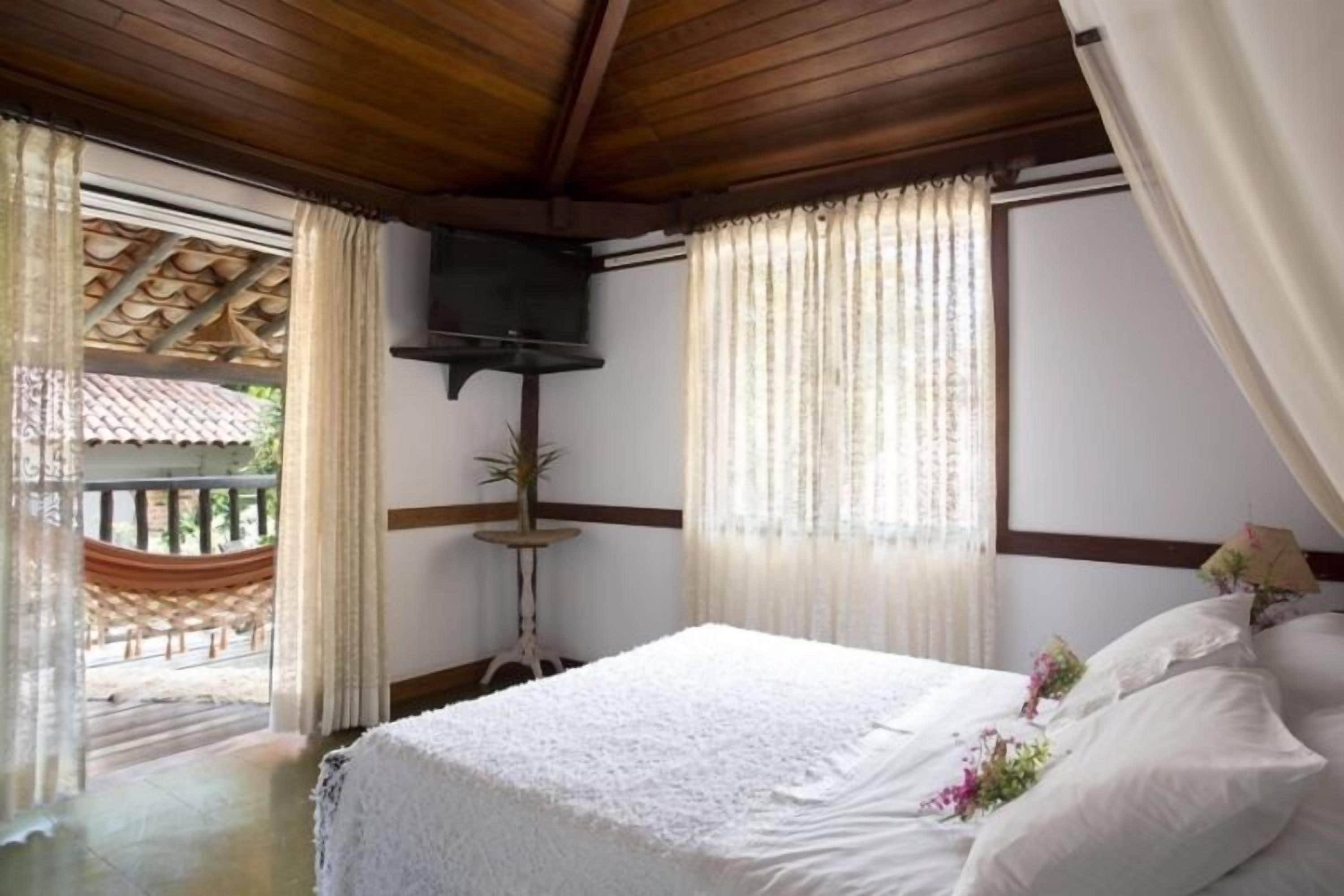 Un dormitorio con techo de madera, una cama con ropa blanca, una mesita con una planta y vista a una hamaca al aire libre.