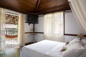 Un dormitorio con techo de madera, una cama con ropa blanca, una mesita con una planta y vista a una hamaca al aire libre.