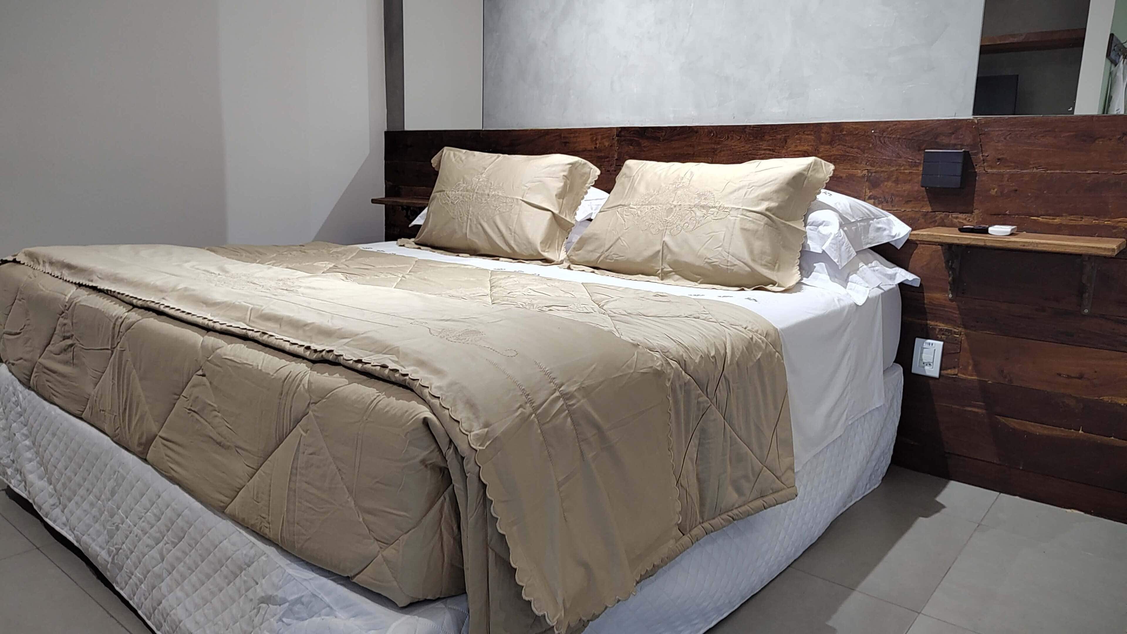 A neatly made bed with a beige quilt and pillows, a wooden headboard, and a bedside table with a lamp.