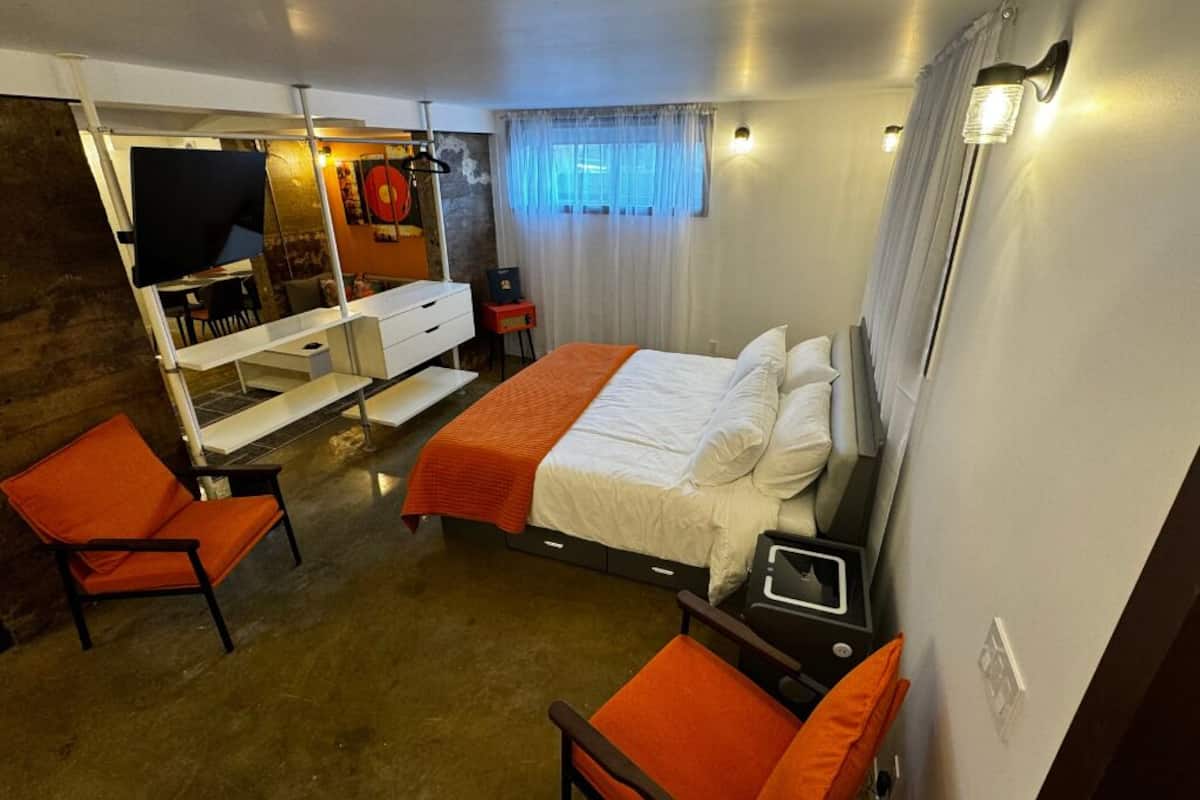 Una habitación de hotel con una cama, dos sillones naranja, un escritorio y un televisor.