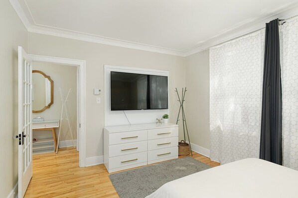 A bedroom with a bed, a white dresser, a television, and a window with curtains.