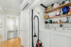 A laundry room with a washing machine, dryer, and a vacuum cleaner. There are shelves with various cleaning supplies and a door leading to another room. - 10/10 Walkscore | Free Parking | Metro (Montreal)