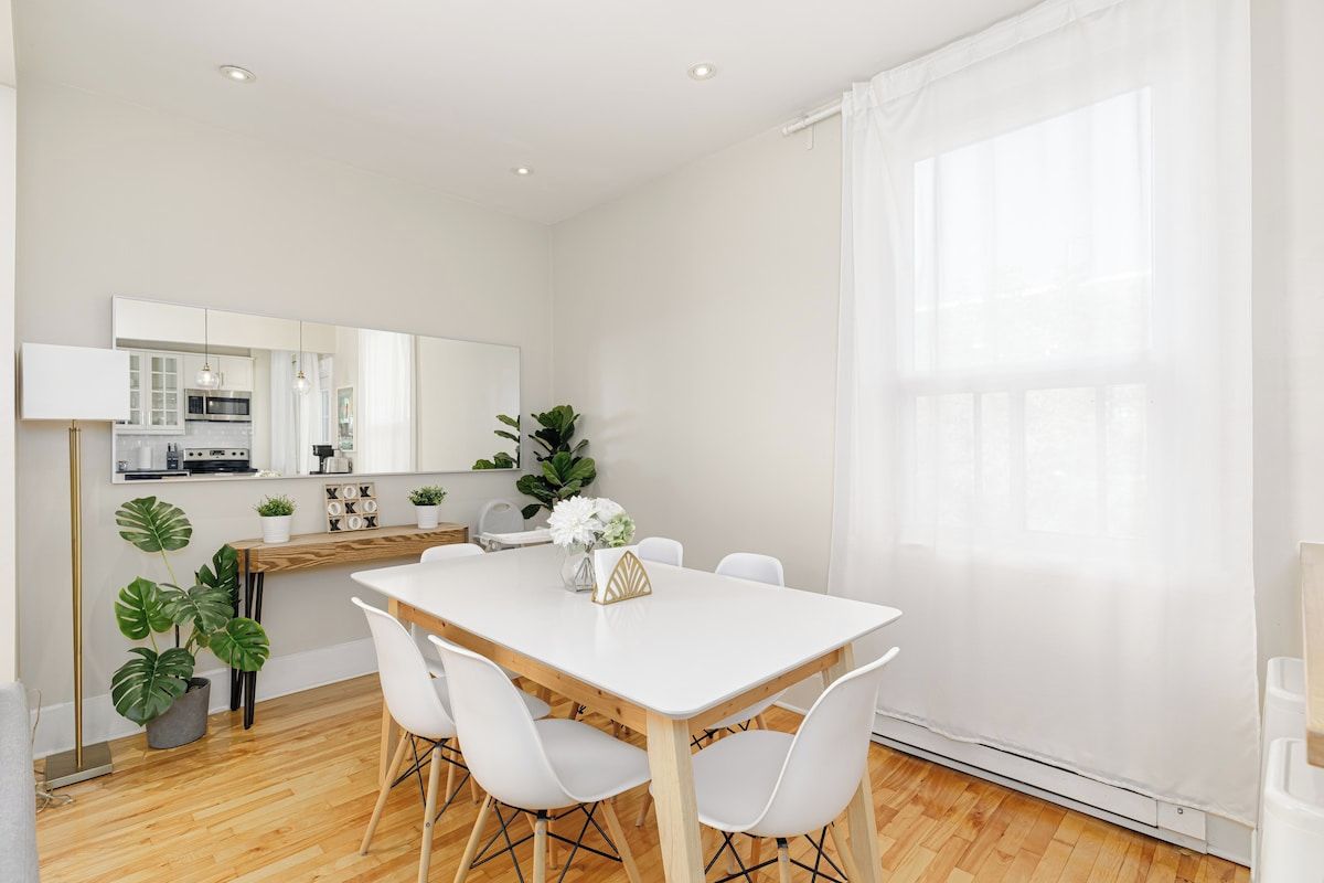 A modern dining area with a white table and chairs, a kitchen in the background, and a large window with sheer curtains.