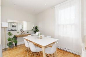 A modern dining area with a white table and chairs, a kitchen in the background, and a large window with sheer curtains.