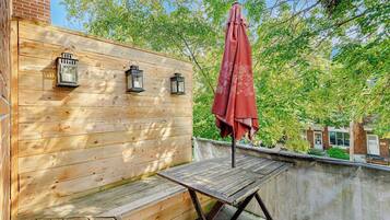 Una terraza de madera con una mesa, sillas, un paraguas rojo y tres faroles fijados a la pared.