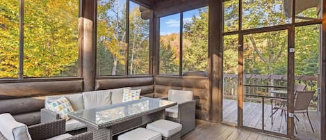 A log cabin with a glass-enclosed porch, wicker furniture, and a view of autumn foliage.