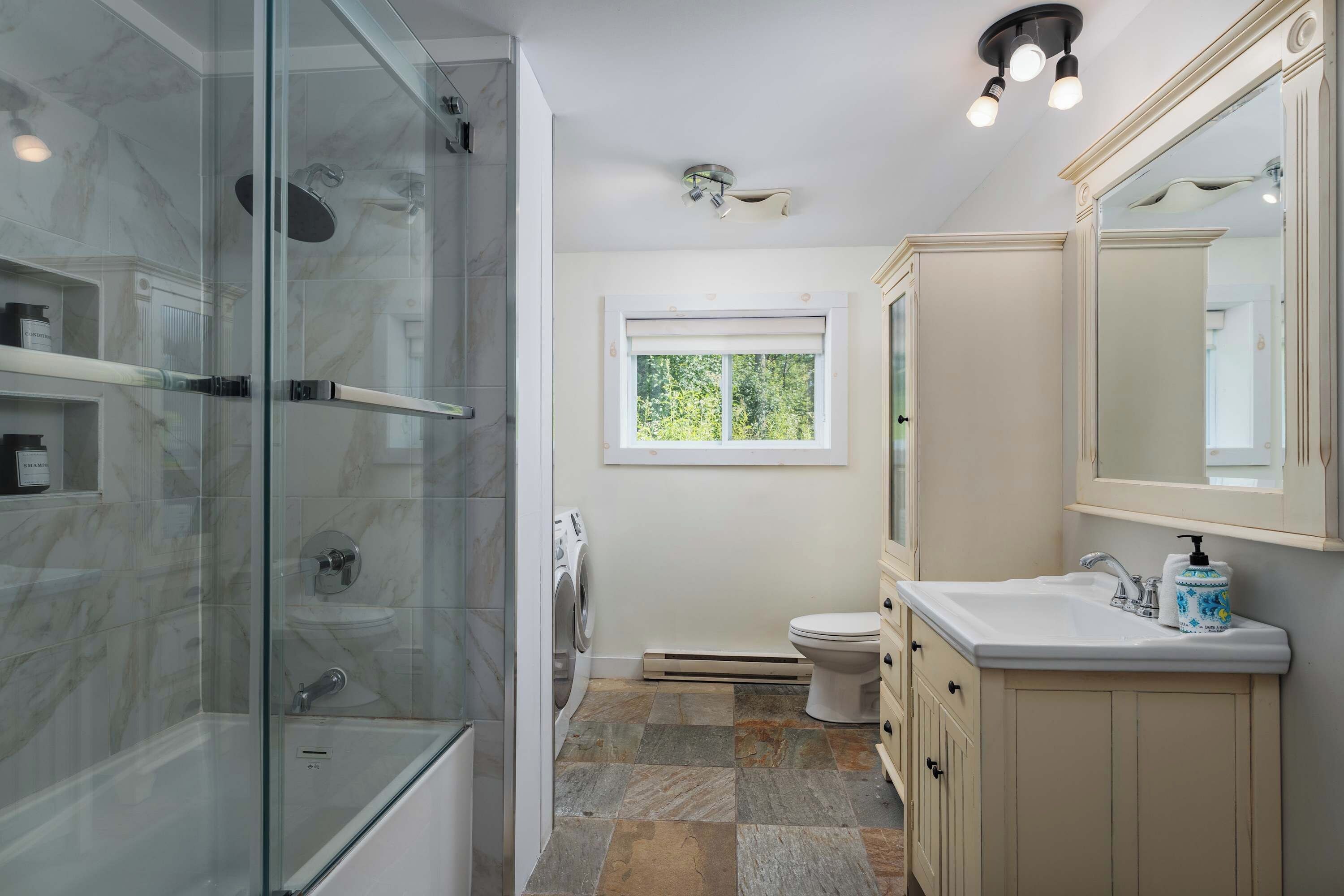 A bathroom with a glass shower enclosure, a toilet, a sink with a mirror, and a window providing natural light.