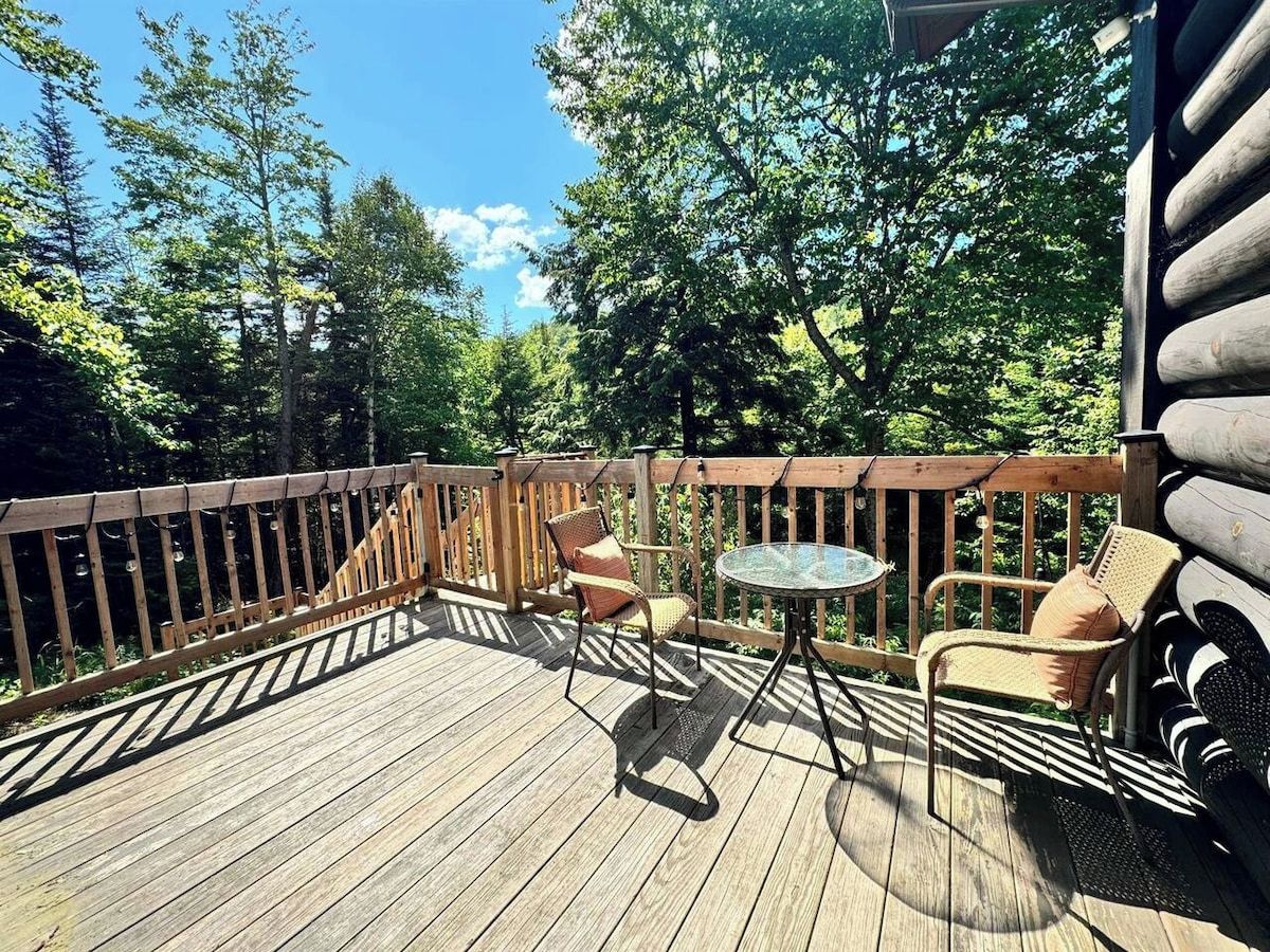 Eine Holzterrasse mit Tisch und zwei Stühlen, umgeben von Bäumen.