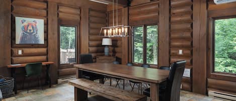A log cabin interior with a large dining table, chairs, a bench, a painting, and a window with a view of trees.