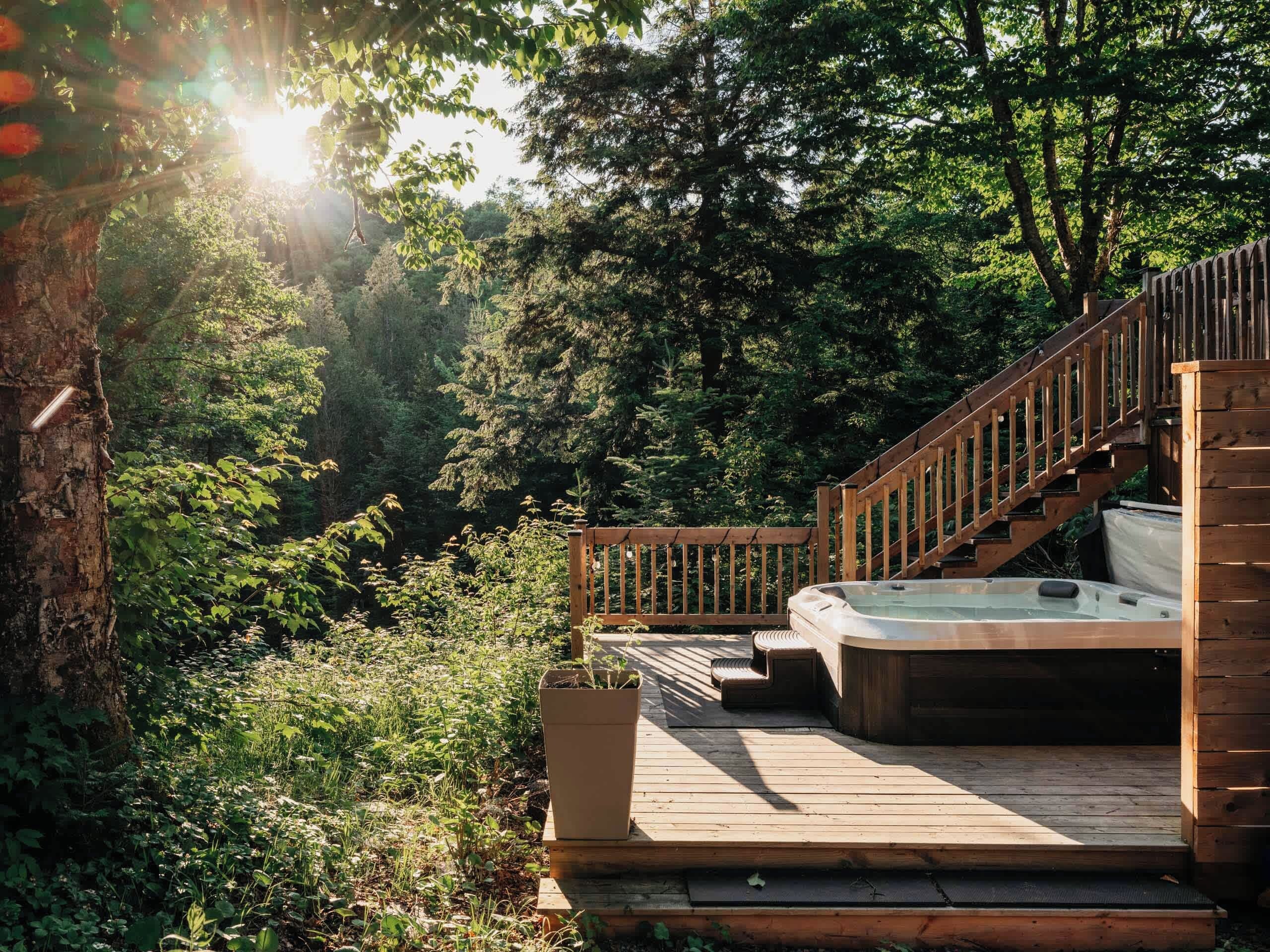 Une terrasse en bois avec un jacuzzi, entourée d’une végétation luxuriante et d’arbres.