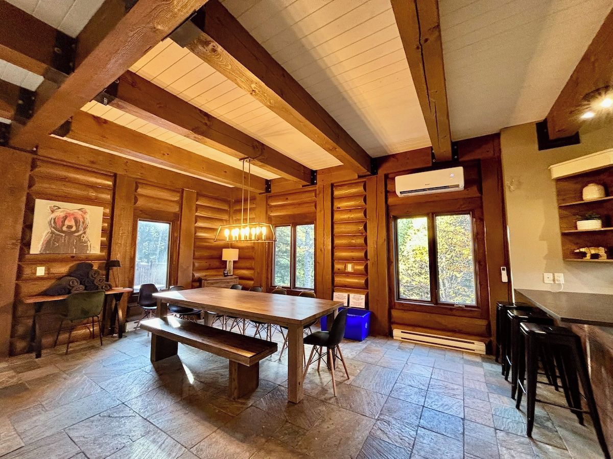A spacious wooden cabin interior with a large dining table, chairs, a bench, a hanging light fixture, and a view through the windows.