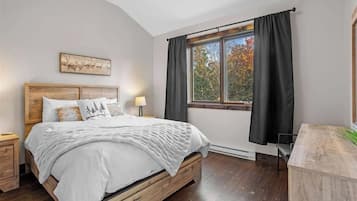 Un dormitorio con cama, mesita de noche, ventana con cortinas y un cuadro en la pared.