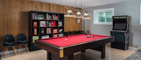 Une table de billard recouverte d’un drap rouge, entourée d’une bibliothèque en bois remplie de jeux de société, de deux chaises noires et d’une autre table de billard recouverte d’un drap rouge.