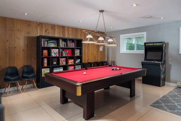 Une table de billard recouverte d’un drap rouge, entourée d’une bibliothèque en bois remplie de jeux de société, de deux chaises noires et d’une autre table de billard recouverte d’un drap rouge.