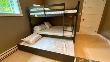 Una cama litera con colchón y almohadas, una ventana con vista a follaje y una puerta.