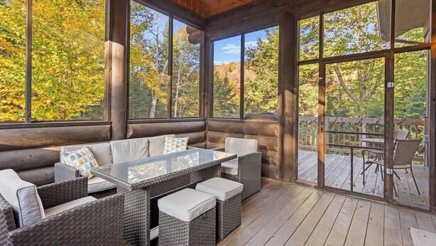 Une cabane en rondins dotée d’une grande porte vitrée, d’un coin repas avec des meubles en osier et d’une vue sur les arbres aux couleurs automnales.