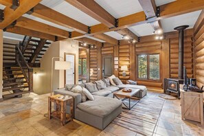 Interior de una cabaña de troncos con escalera, sofá seccional, chimenea y un ventanal grande.