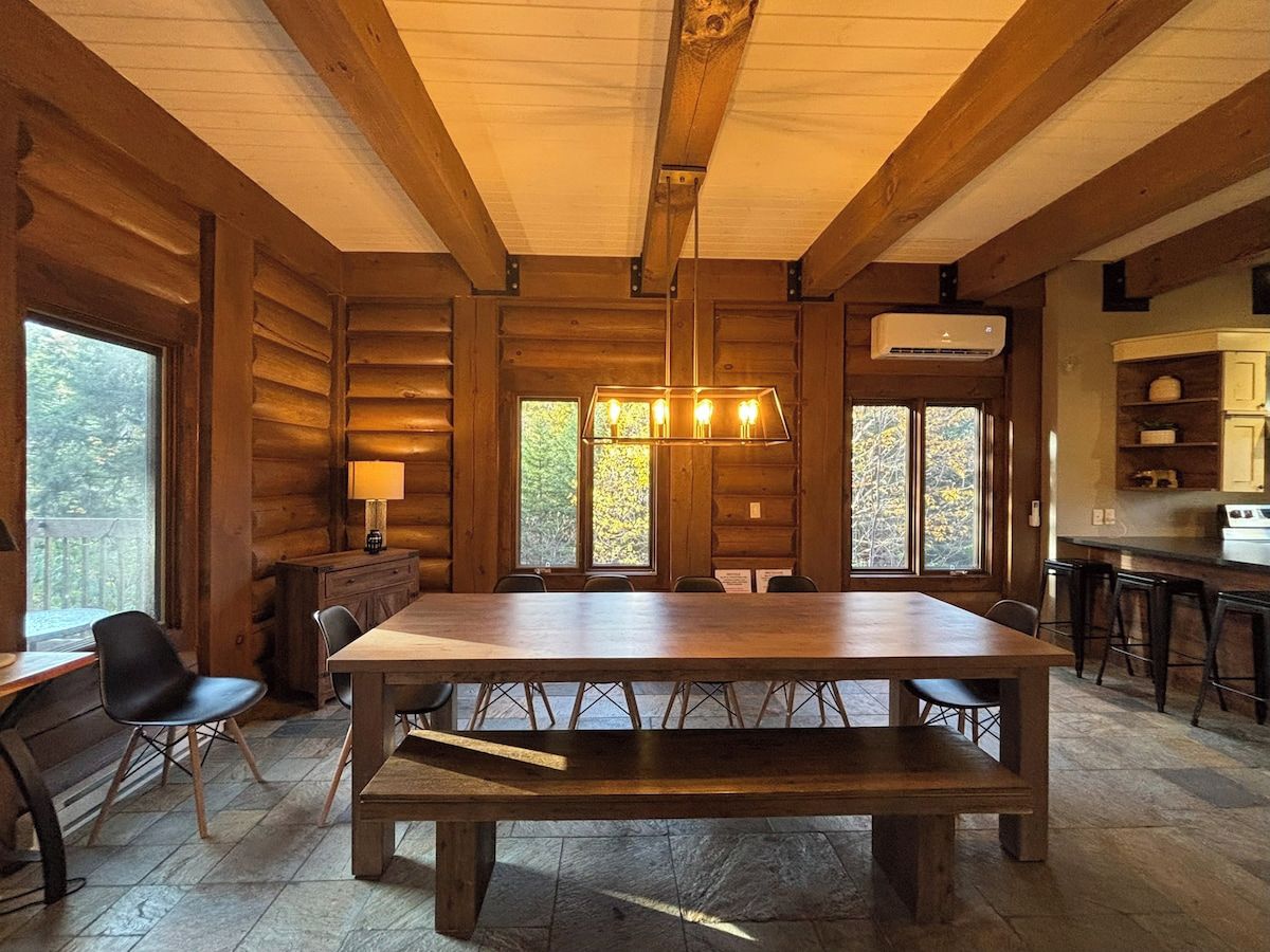 Un intérieur en bois avec une grande table à manger, des chaises, un luminaire suspendu et un coin cuisine équipé de tabourets de bar.
