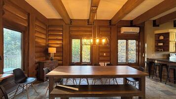Un intérieur en bois avec une grande table à manger, des chaises, un luminaire suspendu et un coin cuisine équipé de tabourets de bar.