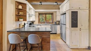 Une cuisine moderne dotée d’un îlot central, de tabourets de bar, d’appareils électroménagers en acier inoxydable et d’un tableau noir sur lequel sont écrites des notes manuscrites.